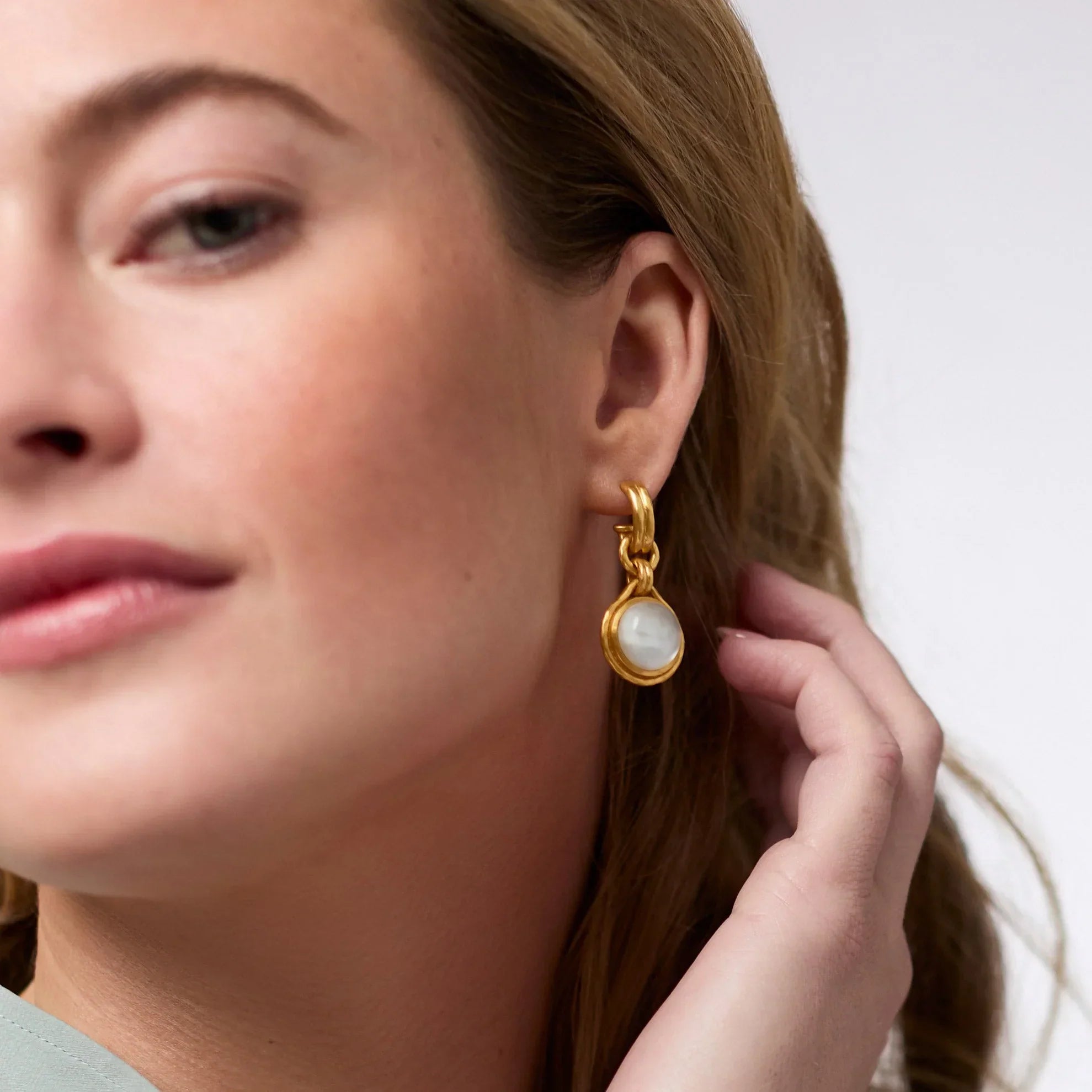 Close-up of a woman wearing a gold earring with a white gemstone on a neutral background