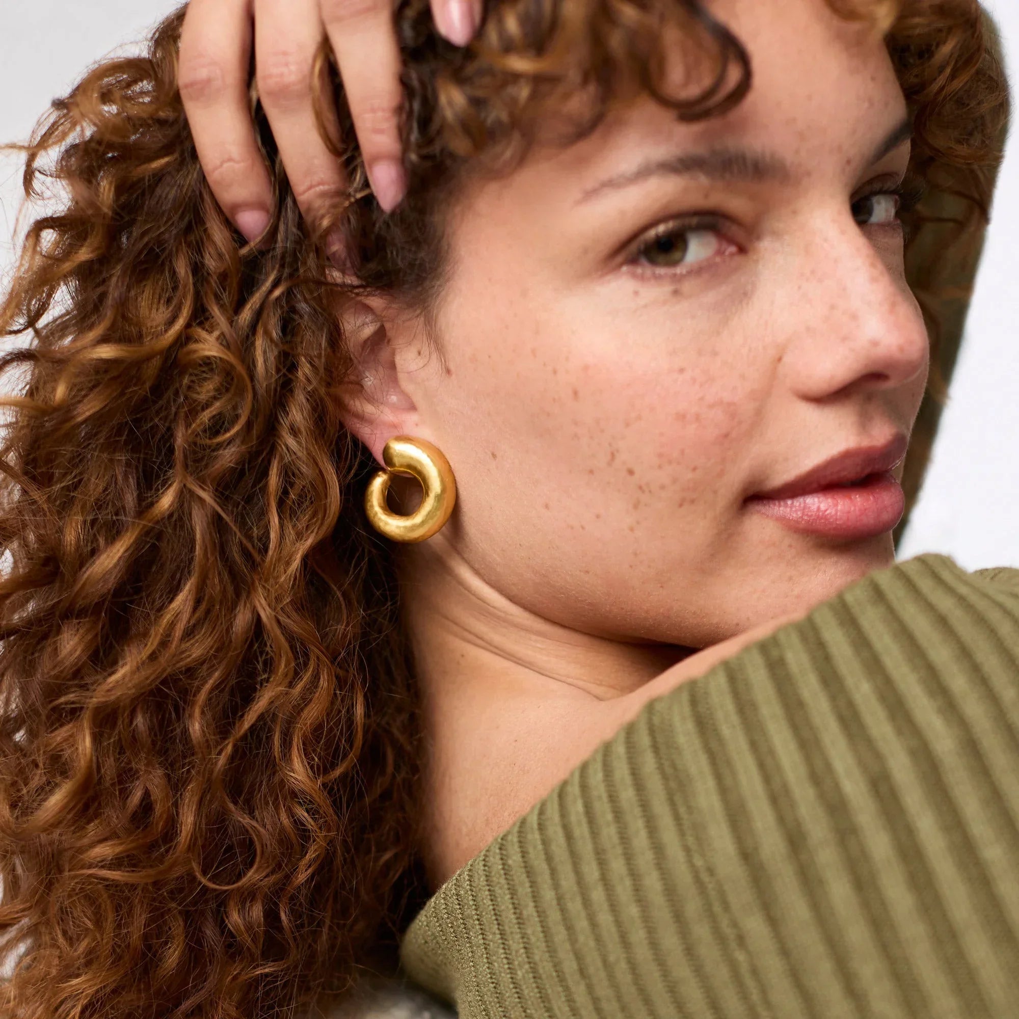 Close-up of a woman with curly hair wearing gold earrings, with a neutral background.